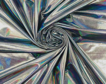 May include: A metallic silver fabric with a high-gloss, holographic finish that reflects a spectrum of rainbow colors. The material is gathered into a central spiral, creating deep, radiating folds and pleats that emphasize the reflective surface.