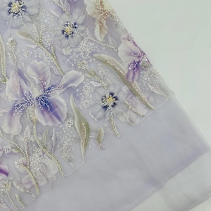 May include: A sheer lavender fabric featuring intricate floral embroidery with purple and white petals. The design includes delicate beadwork and sequins accentuating the flower centres and stems, creating a textured, layered appearance on the mesh.