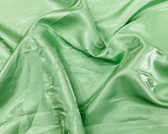 May include: A close-up of a light green fabric with a shiny, silky texture. The fabric is draped in soft folds, creating shadows and highlights. The overall appearance is smooth and flowing, with a subtle sheen.