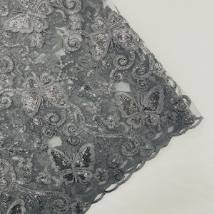 May include: Close-up of a gray fabric with an intricate butterfly and floral design. The fabric is embellished with sequins, creating a shimmering effect. The edges of the fabric have a scalloped trim.