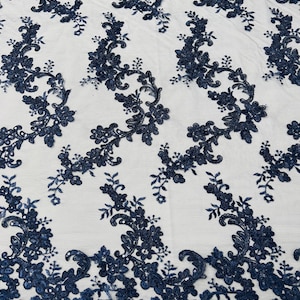 May include: Navy blue floral lace fabric. The fabric features an intricate pattern of embroidered flowers and leaves. The design is detailed with sequins and a scalloped edge. The lace is set against a white background.