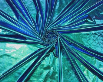 May include: A metallic fabric featuring a vibrant gradient of deep blue and bright teal. The material is gathered in a central spiral, creating radiating folds that emphasize the reflective, high-gloss surface and the shifting color tones.