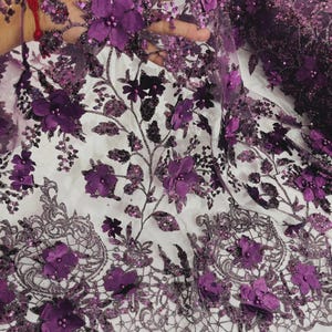 May include: A sheer fabric with a floral design. The fabric features 3D purple flowers, sequins, and embroidered details. The design includes a scalloped edge with floral and lace patterns. The background is white.