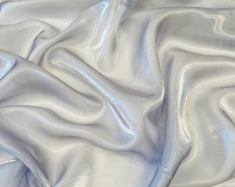 Może przedstawiać: Zbliżenie jasnoszarej, połyskującej tkaniny o płynnym, drapowanym wyglądzie. Tkanina ma miękki połysk i wydaje się lekka, z subtelnymi odcieniami tworzącymi głębię i fakturę. Fałdy i krzywizny materiału odbijają światło.