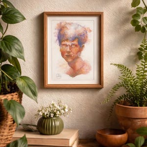 May include: A framed watercolor portrait of a person with orange and blue hair. The artwork is in a wooden frame and hangs on a neutral-colored wall. The portrait is part of a home decor display with plants and books.