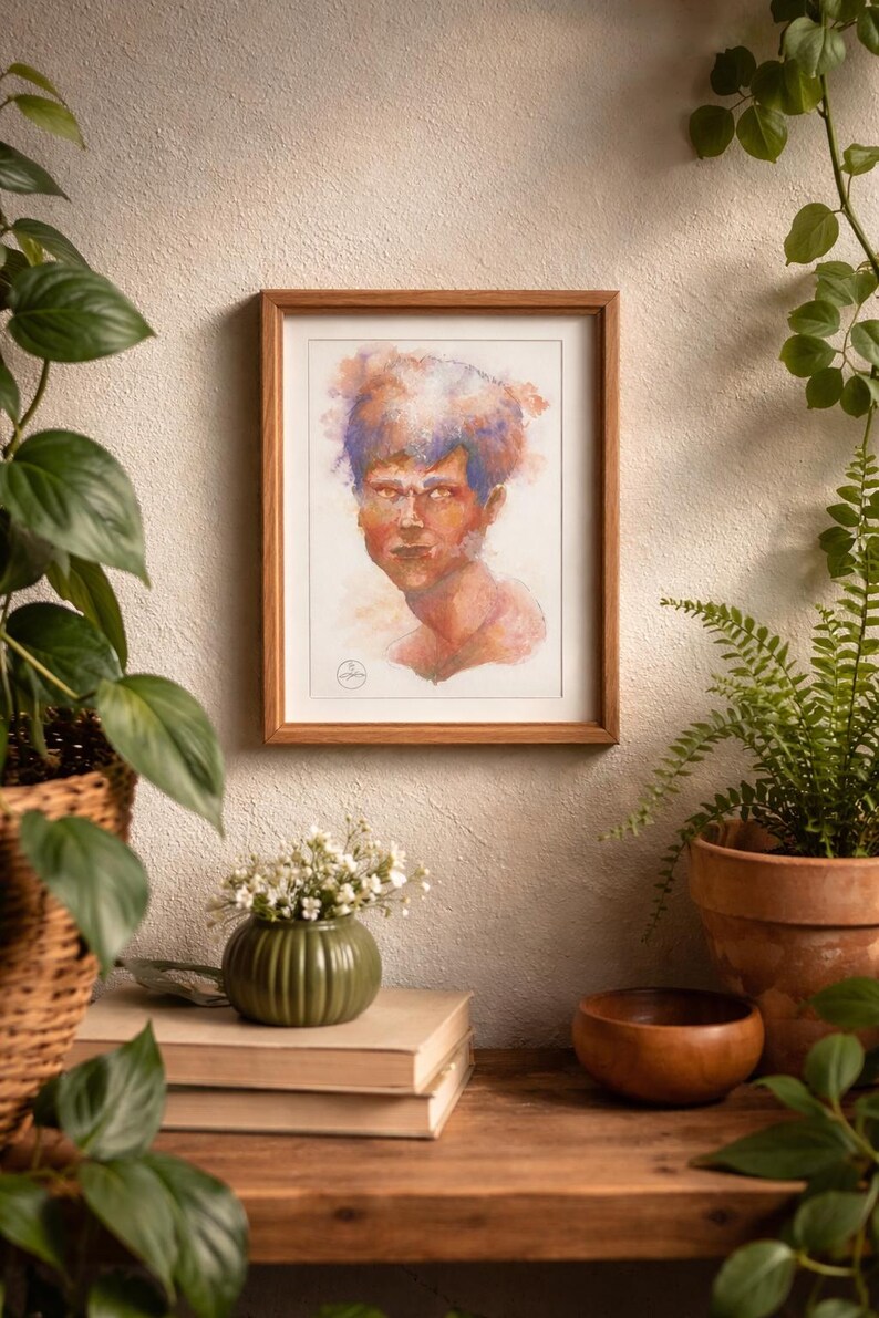 May include: A framed watercolor portrait of a person with orange and blue hair. The artwork is in a wooden frame and hangs on a neutral-colored wall. The portrait is part of a home decor display with plants and books.