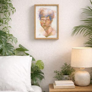 May include: A framed watercolor portrait of a person with blue and orange hair. The artwork is displayed on a white wall, surrounded by greenery and a bedside table with a lamp and books.