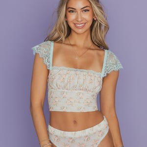 May include: A light blue cropped top and matching bottoms set featuring a delicate floral pattern in shades of pink and green. The top has a square neckline, gathered fabric detailing, and short cap sleeves made of intricate light blue lace.