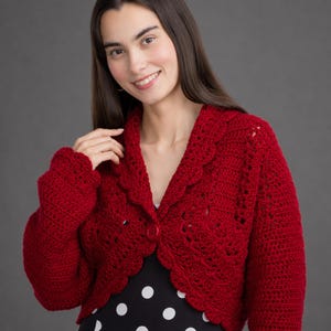 May include: A deep red crochet bolero jacket featuring long sleeves, a scalloped edge collar, and a single matching red button closure at the center front. The garment is crafted with a textured stitch pattern and is worn over a black polka dot top.