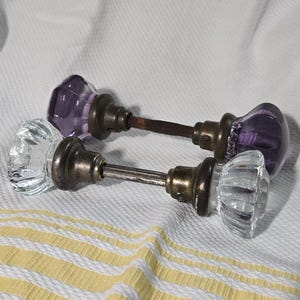 Könnte beinhalten: Zwei Türknäufe mit facettierten Glasknöpfen und Metallbasen, die durch einen zentralen Dorn verbunden sind. Ein Set hat klares Glas, das andere violettes Glas. Beide Sets verfügen über Metallkomponenten in einer gealterten Messingoptik.