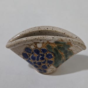 以下が含まれることがあります： 斑点のあるベージュの釉薬が施された、扇形の小さな陶器製容器。前面には、青いブドウと緑の葉が手描きされており、金色のアクセントが加えられています。開口部は幅広く湾曲しており、底部に向かって細くなっています。