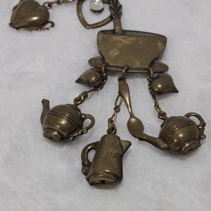 Könnte beinhalten: Eine Brosche aus messingfarbenem Metall mit einer zentralen Teetassenform und mehreren Anhängern, die an kleinen Ketten befestigt sind. Die Anhänger umfassen zwei Teekannen, eine Kaffeekanne, zwei Löffel, zwei kleine Herzen und einen herzförmigen Rahmen mit einem klaren Stein.