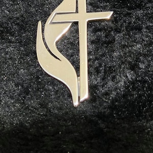 May include: A silver-toned metal pendant featuring the United Methodist Church emblem, which consists of a cross positioned next to a stylized flame. The pendant is attached to a silver-toned chain link necklace with a circular jump ring connector.