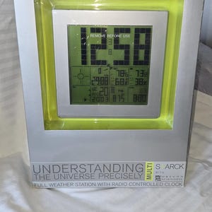 May include: A silver and lime green digital weather station in its original packaging. The device features a square screen displaying the time 12:58, date, and various weather metrics. The box includes the text "UNDERSTANDING THE UNIVERSE PRECISELY" and "MULTI STARCK WITH OREGON SCIENTIFIC".