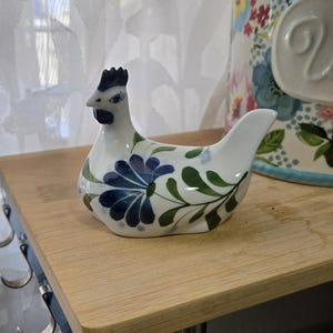 May include: A white ceramic chicken figurine featuring a hand-painted blue floral pattern on its body, a dark blue comb, and a dark blue wattle. The chicken is depicted in a seated position with its head turned slightly to the left side.