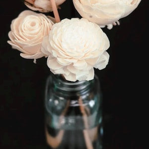 May include: A clear glass bottle containing four cream-colored wooden flowers with layered petals. The flowers are attached to thin, light brown wooden stems that extend into the bottle. The arrangement features three fully bloomed flowers and one smaller, tighter bud, all set against a solid black background.