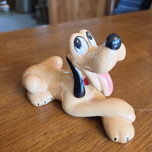 May include: A ceramic figurine of a light tan dog with long black ears, a black nose, and a pink tongue hanging out. The dog has large white eyes with black pupils and a small red collar detail. The surface of the figurine has a glossy finish.