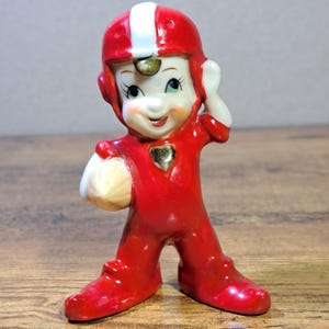 May include: A vintage ceramic figurine of a football player in a red uniform and helmet with a white stripe. The figurine is holding a football and has a gold heart on the chest. The figure has a smiling face and is standing on a wooden surface.