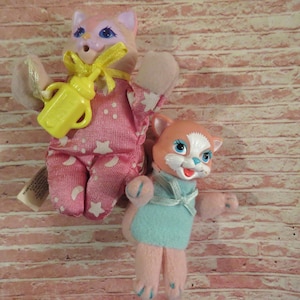 May include: Two plush pink and white cat toys. One cat is wearing pajamas with stars and holding a yellow bottle. The other cat is wearing a blue dress.