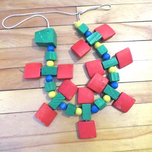 May include: A colorful wooden toy with red, green, yellow, and blue components. The toy is made of square and cube-shaped blocks and round beads, connected by a white string. The toy is on a wooden surface.