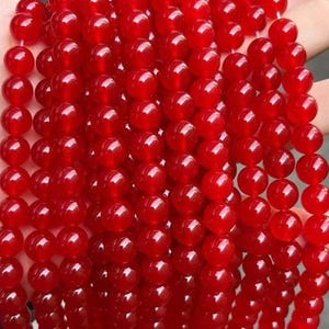 Puede incluir: Una colección de múltiples hilos de cuentas rojas, lisas, redondas y vibrantes. Cada cuenta presenta un acabado pulido y brillante que refleja la luz, creando una apariencia uniforme en las numerosas cuerdas verticales de componentes.