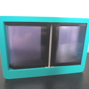 May include: A teal-colored picture frame with a rounded rectangular shape. The frame holds two black-framed panels with a purple hue. The frame is sitting on a dark surface.