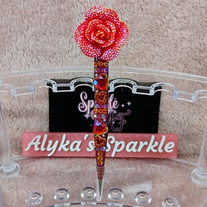 May include: A decorative pen with a red, rhinestone-covered rose at the top. The pen's shaft is adorned with red heart-shaped gems and rhinestones. The pen is set against a background with the text "Alyka's Sparkle".