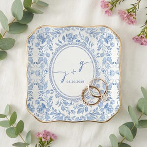 May include: Square, floral-patterned ring dish with a gold rim. The dish features a blue and white floral design and a central circle with the initials "J + g" and the date "03.20.2025". Two gold rings rest on the dish.