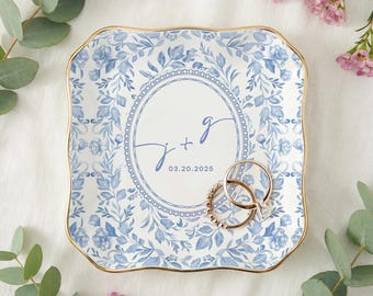 May include: Square, floral-patterned ring dish with a gold rim. The dish features a blue and white floral design and a central circle with the initials "J + g" and the date "03.20.2025". Two gold rings rest on the dish.