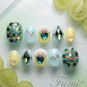 Puede incluir: Un conjunto de diez uñas postizas con una paleta de colores pastel en azul claro y amarillo pálido. Los diseños incluyen purpurina verde e iridiscente, pedrería en forma de corazón, detalles de perlas y gemas verdes en forma de rombo.