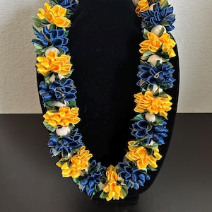 May include: A vibrant lei necklace with alternating clusters of yellow and navy blue satin flowers, accented with white spherical beads and green leaves. The lei is displayed on a black mannequin, showcasing its colorful design.