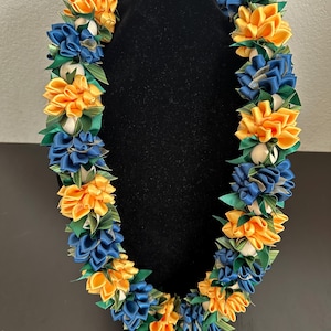 May include: A vibrant lei necklace featuring alternating clusters of yellow and navy blue satin ribbon flowers, accented with green leaves and white accents. The lei is displayed on a black mannequin head, showcasing its intricate design and color contrast.