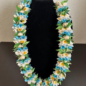 May include: A lei necklace with white, yellow, and blue ribbon flowers and green leaves. The lei is displayed on a black mannequin head. The flowers are interspersed with small, round, light-colored beads.