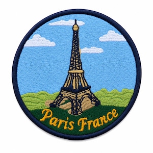 May include: A circular embroidered patch featuring the Eiffel Tower in tan and dark blue thread against a light blue sky with white clouds and green hills. The text "Paris France" is stitched in yellow cursive at the bottom of the patch.