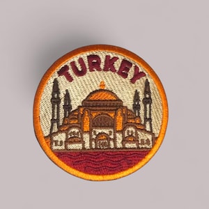 Può includere: Toppa rotonda ricamata con la scritta "TURKEY" in alto. La toppa raffigura un'illustrazione dettagliata di un edificio con una cupola e minareti. Ha uno sfondo beige, bordo arancione e dettagli rossi.