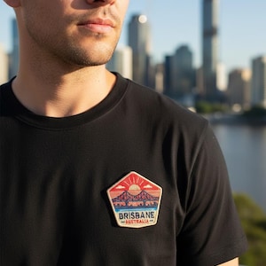 May include: Black crew neck t-shirt with a Brisbane, Australia patch. The patch features a bridge silhouette under a setting sun, with the city skyline in the background. The patch has red, orange, blue, and white colors.