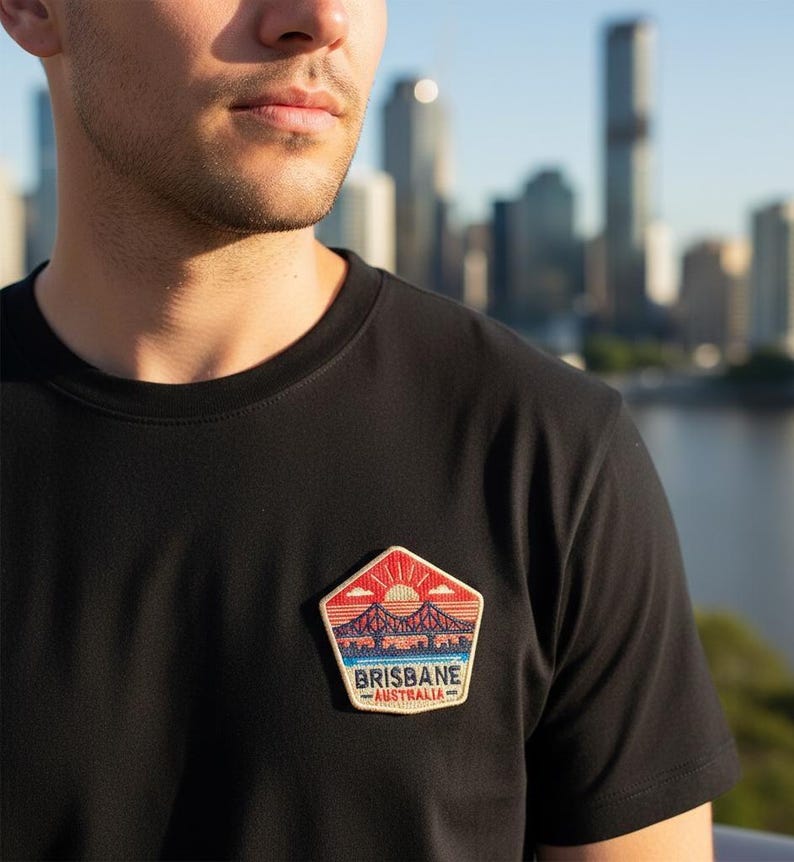 May include: Black crew neck t-shirt with a Brisbane, Australia patch. The patch features a bridge silhouette under a setting sun, with the city skyline in the background. The patch has red, orange, blue, and white colors.