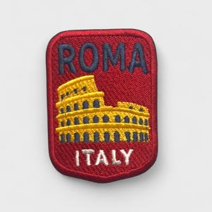 Puede incluir: Un parche rectangular rojo con la palabra "ROMA" en azul marino, sobre una ilustración amarilla y azul marino del Coliseo. La palabra "ITALY" está bordada en blanco debajo de la ilustración.