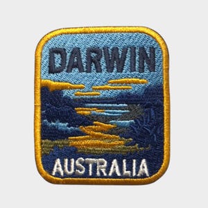 May include: Embroidered patch featuring the word "DARWIN" above "AUSTRALIA". The patch has a blue and yellow landscape design with a gold border. The design includes a river or waterway.