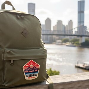May include: Olive green backpack with a front pocket and a patch that reads "Brisbane Australia" with a bridge and sunset design. The backpack has a diamond-shaped leather tag and two zippered compartments. The background shows a city skyline and a river.