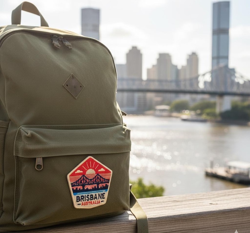 May include: Olive green backpack with a front pocket and a patch that reads "Brisbane Australia" with a bridge and sunset design. The backpack has a diamond-shaped leather tag and two zippered compartments. The background shows a city skyline and a river.