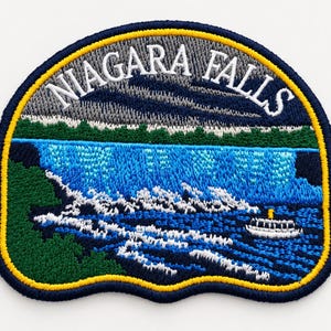 May include: An embroidered patch featuring a landscape of Niagara Falls with a blue waterfall, green trees, and a small white boat. The text NIAGARA FALLS is stitched in white at the top against a dark blue sky. The patch has a dark blue border.