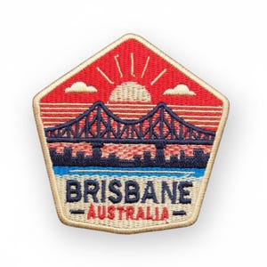 Puede incluir: Parche bordado con un puente sobre el agua y un diseño de puesta de sol. El parche tiene forma de pentágono y las palabras "BRISBANE AUSTRALIA" en la parte inferior. Los colores son rojo, azul y beige.