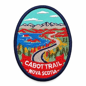May include: An oval embroidered patch featuring a landscape of the Cabot Trail in Nova Scotia. The design includes a winding gray road, blue water, green and red trees, and gray mountains under a light blue sky. A red banner displays the text CABOT TRAIL NOVA SCOTIA.