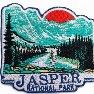May include: Embroidered patch depicting a landscape of Jasper National Park. The design shows mountains, a river, and a red sun. The text "JASPER NATIONAL PARK" is embroidered in blue on a white background, with a blue border.