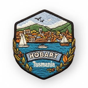 May include: Embroidered patch featuring a scenic view of Hobart, Tasmania. The design includes a harbor with sailboats, buildings, and mountains under a blue sky. The word "HOBART" is prominently displayed, with "Tasmania" below.