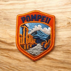 Può includere: Toppa ricamata con la scritta "Pompei" in blu su uno scudo arancione. La toppa raffigura un paesaggio con rovine, una montagna e un fiume, con colori blu, arancione e bianco.