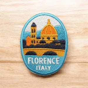 Może przedstawiać: Okrągła, haftowana naszywka z panoramą Florencji. Projekt przedstawia kopułę Duomo, dzwonnicę i most w odcieniach pomarańczu, żółci i czerni, na niebieskim tle. U dołu naszywki wyhaftowano napis "FLORENCE ITALY".