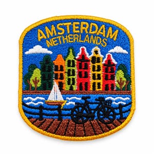 May include: An embroidered patch featuring a blue sky with white clouds above colourful canal houses. The text AMSTERDAM NETHERLANDS is stitched in yellow. Below, a white sailboat floats on blue water near a black bicycle on a brown dock.