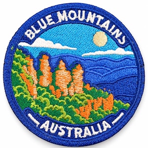 May include: A circular embroidered patch featuring a landscape of the Blue Mountains in Australia. The design includes orange rock formations, green foliage, a blue sky with white clouds, and a yellow sun. The text reads Blue Mountains Australia.